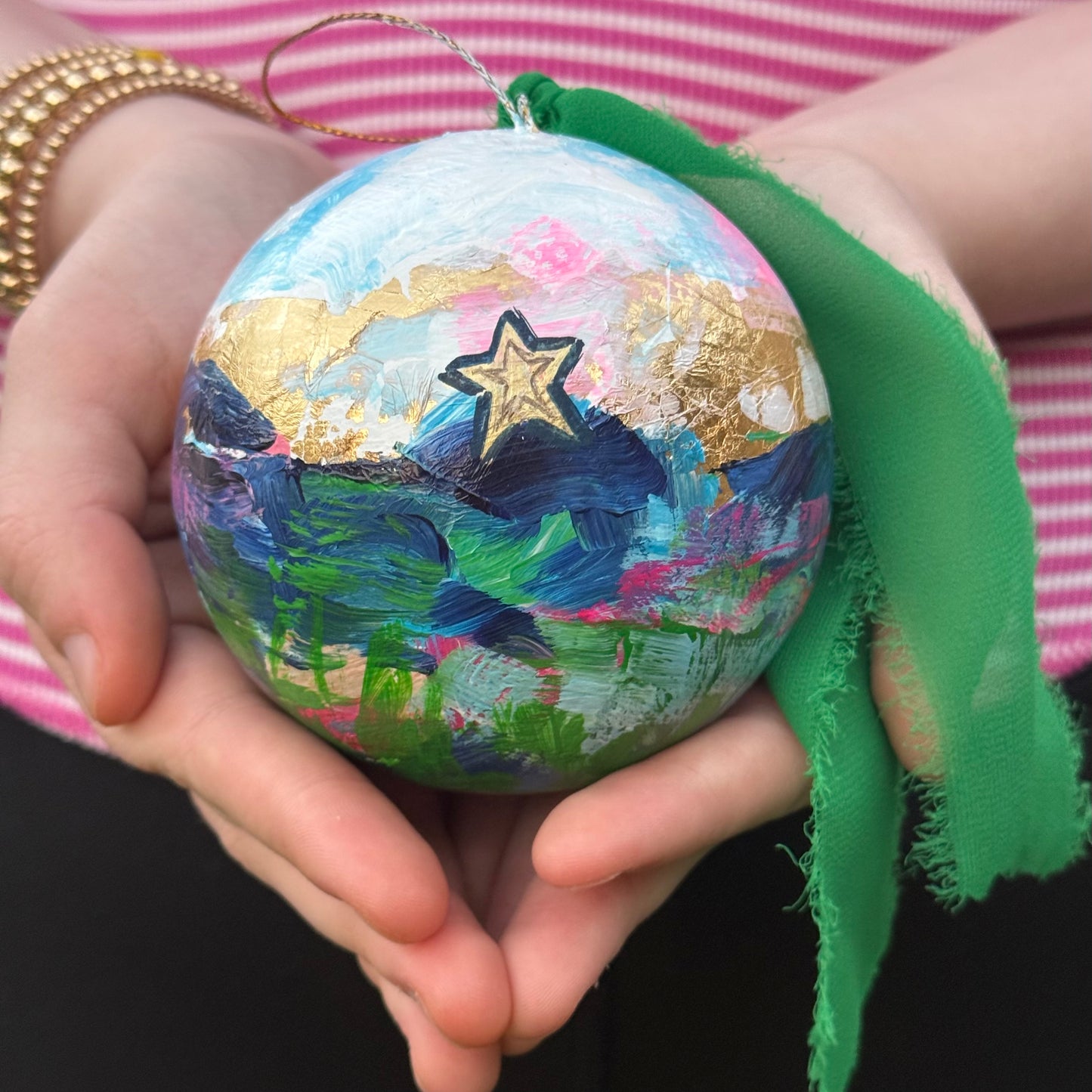 A Colorful painted Christmas ornament by artist Julie Hamilton of the Mill Mountain Star in Roanoke held in 2 hands with a green ribbon, against a striped fabric background.