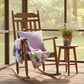 Wooden rocking chair with a colorful blanket and pillow on a wooden deck with a view of trees.