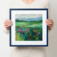 Person holding a framed painting of a field with flowers and mountains.