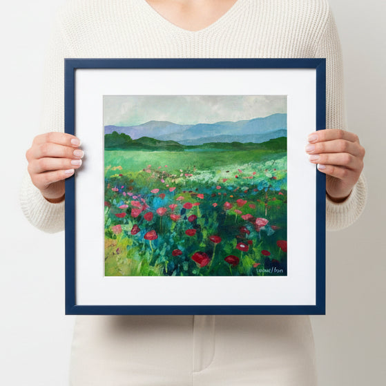 Person holding a framed painting of a field with flowers and mountains.