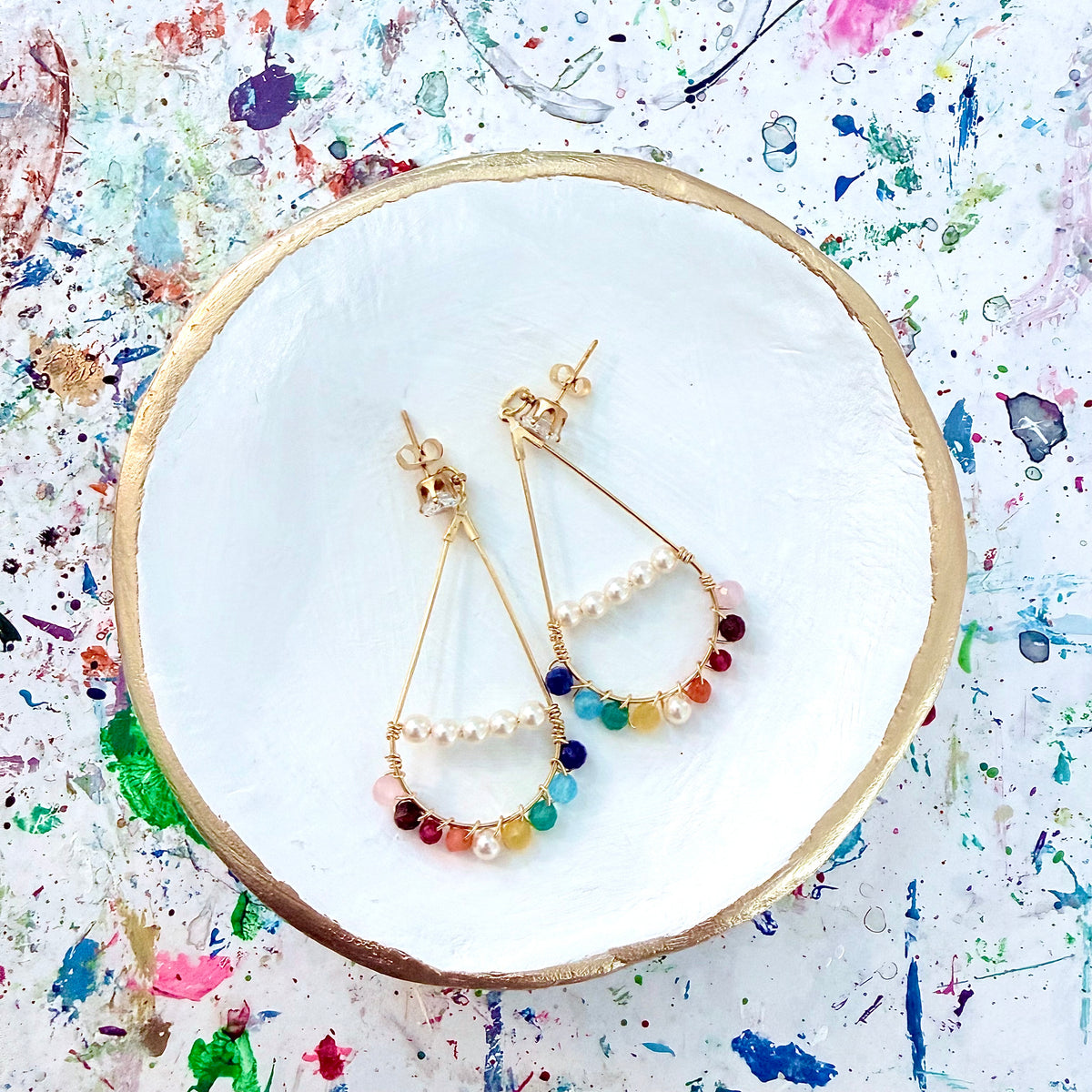 Pair of gold earrings with colorful beads on a white plate against a multicolored splatter background