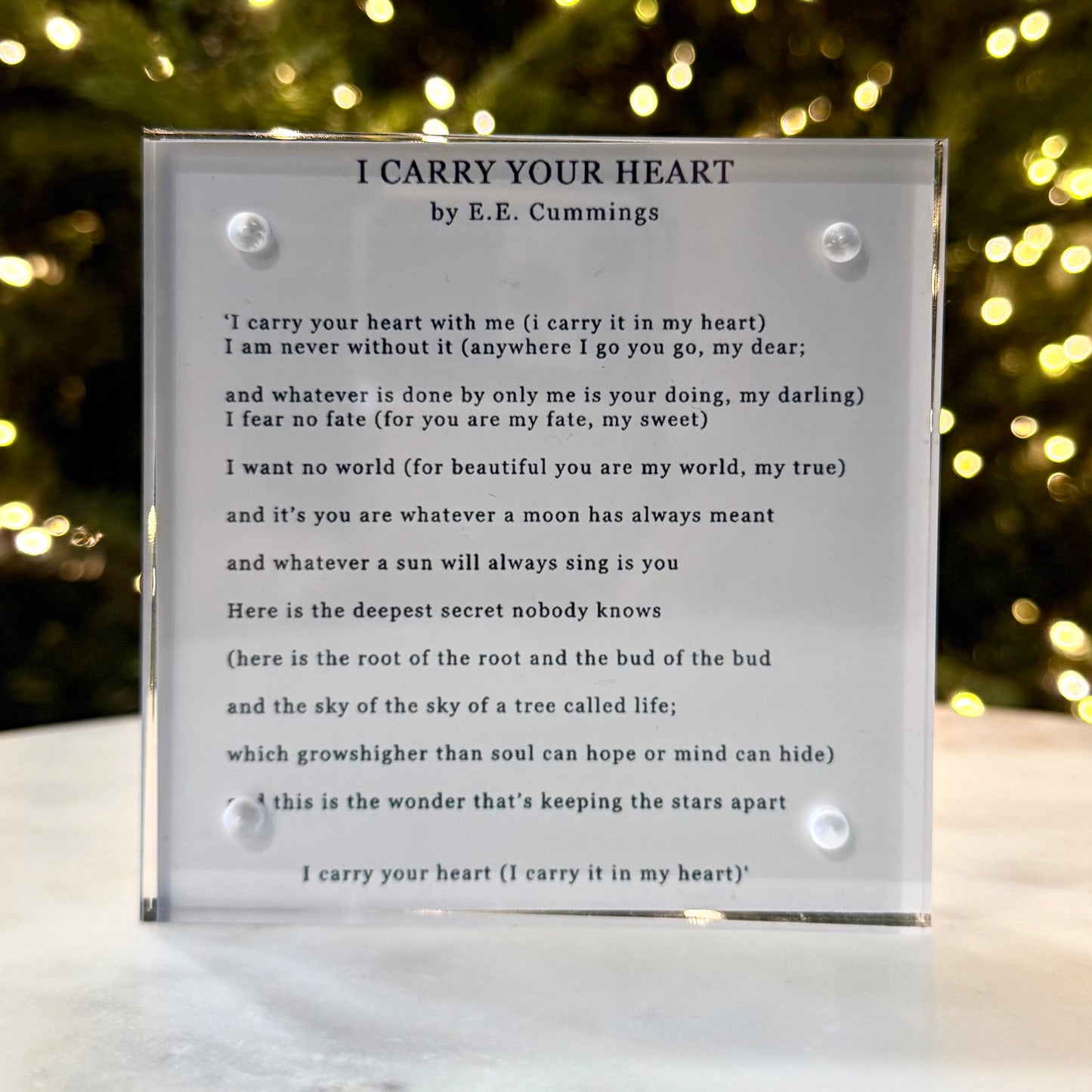 Acrylic plaque with poem 'I Carry Your Heart' by E.E. Cummings against a blurred festive background.