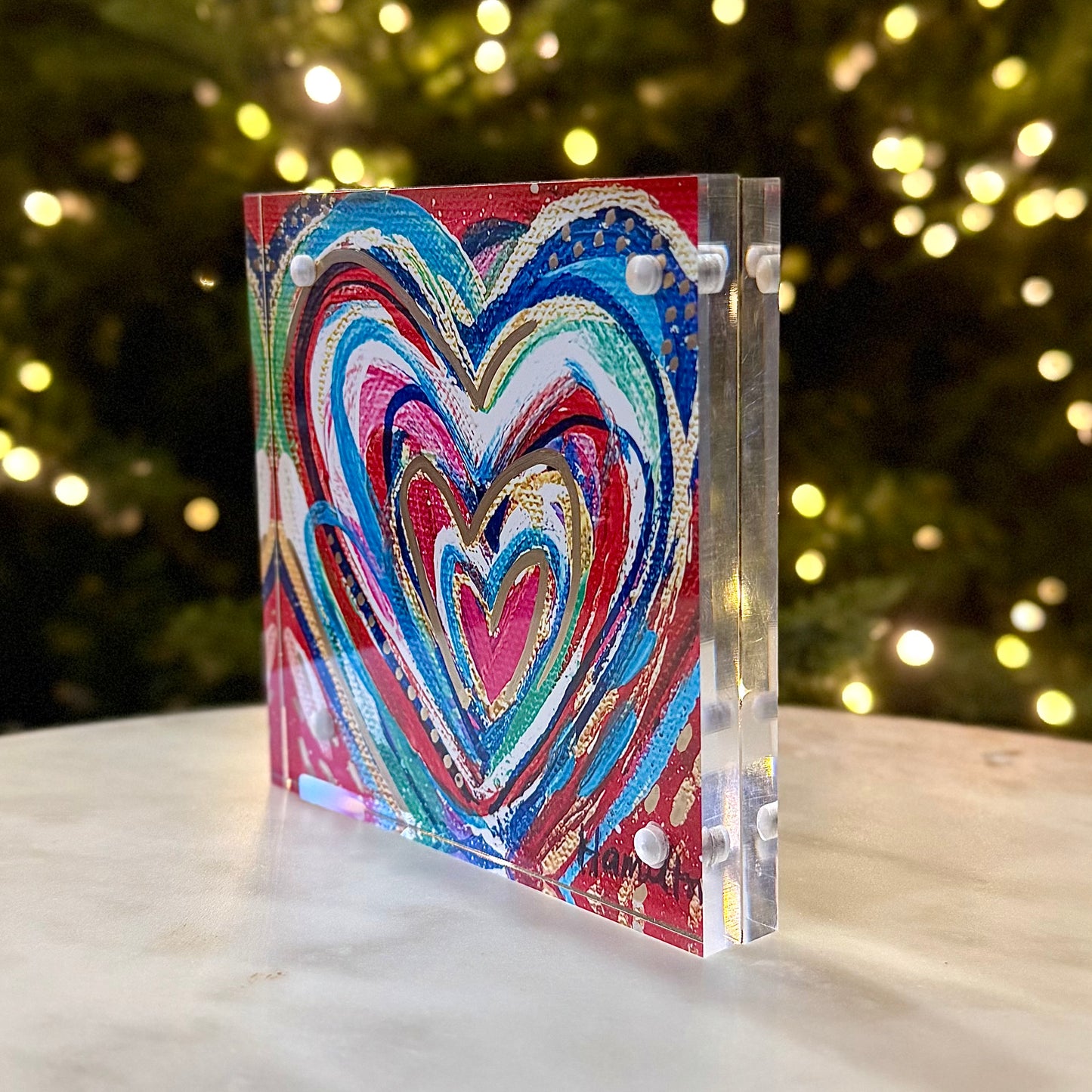 Colorful heart-shaped artwork on a clear stand with a blurred festive background