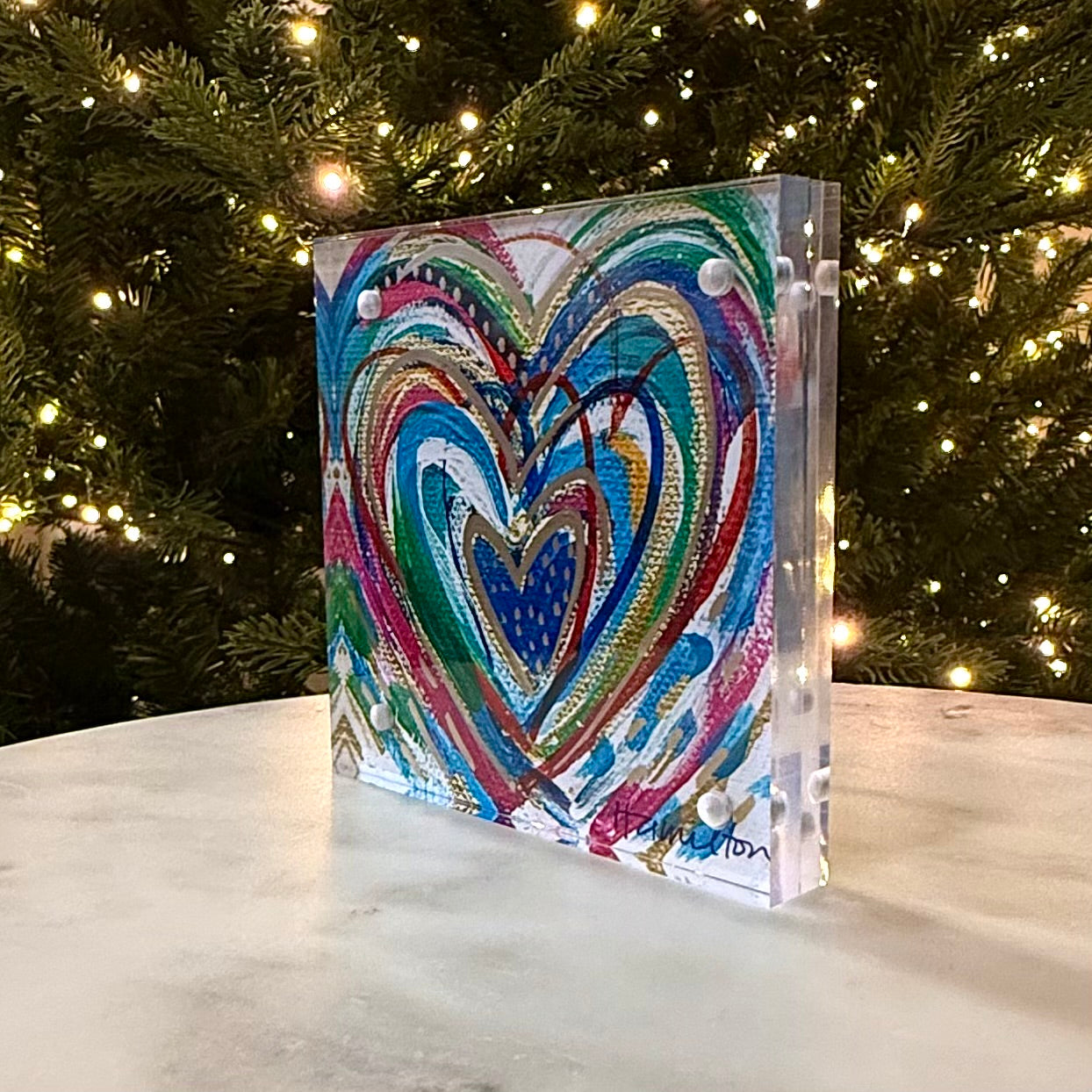 Decorative heart-shaped object with colorful design on a marble surface with Christmas tree in the background