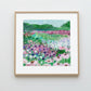 Framed artwork of a colorful floral landscape with mountains on a white wall.
