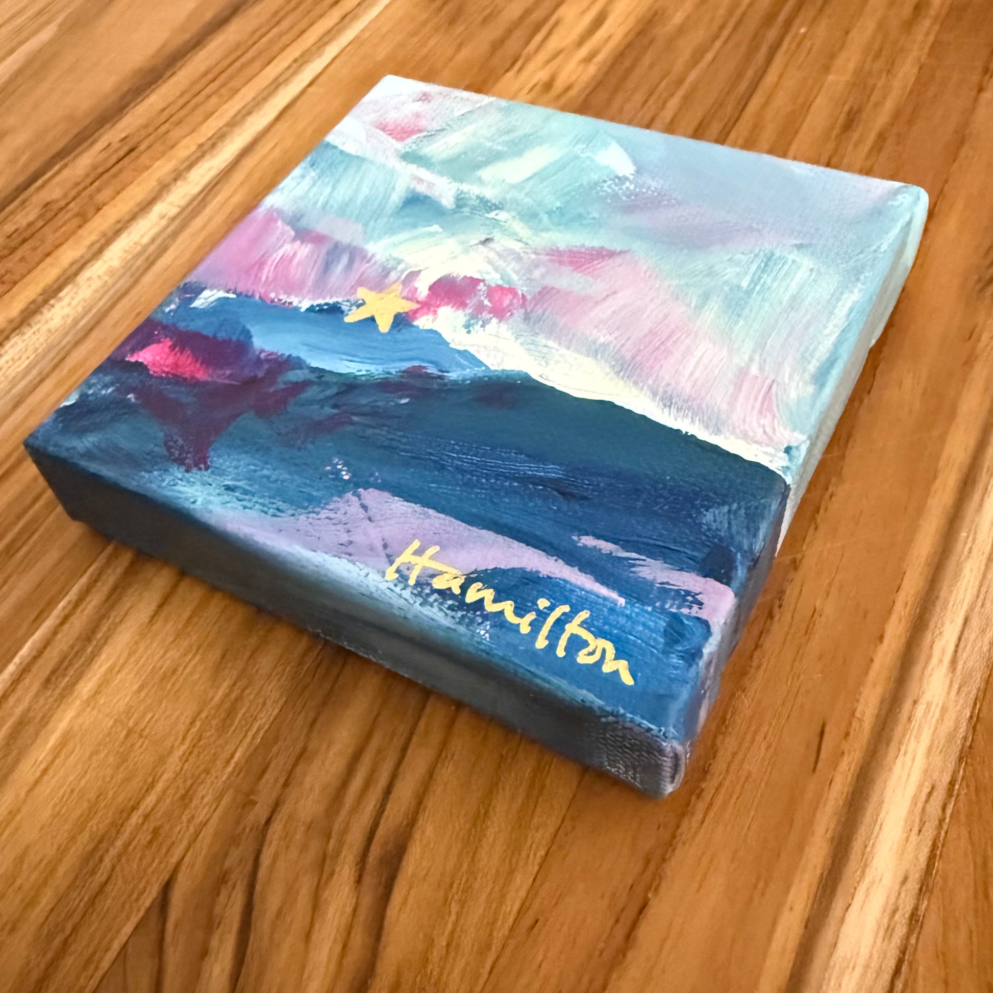 Small square canvas with a colorful abstract mountain landscape of mill mountain star signed by 'Hamilton' on a wooden surface
